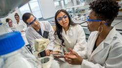 三名CELS学生戴着实验室安全眼镜，穿着外套，在显微镜前交谈